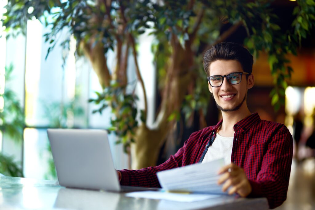 Smiling young hipster man in glasses with laptop on the table. Programmer, web developer.